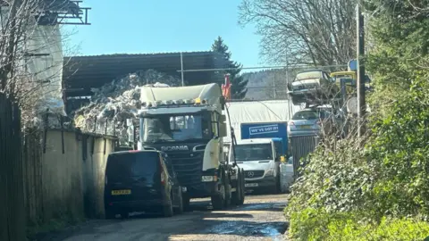 A narrow lane is next to an industrial site with several vans and a large lorry nearby. A large pile of mixed waste and rubble is stacked behind a metal fence, with an excavator visible among the debris and trees in the background on a bright sunny day.