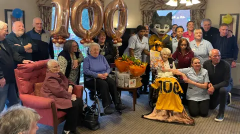 Avery Healthcare Group Joan is sat in a chair with a Wolves shirt draped over her lap and she is holding a birthday card. She is surrounded by former players, club associates, care home staff and friends.