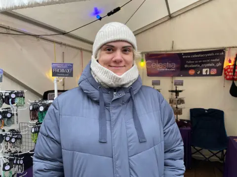 Emily stands in front of her stall in a blue padded jacket, hat and scarf. Her stall selling jewellery is in the background.