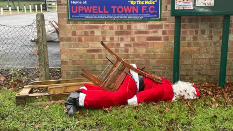 Norfolkinsunshine/BBC Weather Watchers A Father Christmas on a wooden chair was sat outside a football club next to a sign and in front of a wall. However, the figure has fallen over on its side due to the wind.