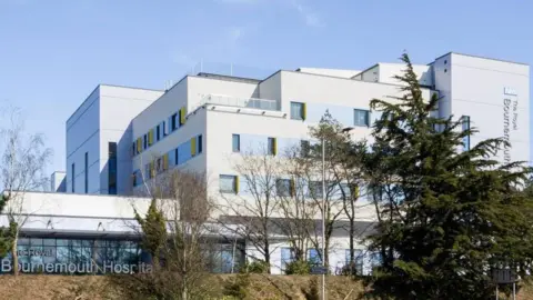 The Beach building at Royal Bournemouth Hospital, as seen from distance. The white, compartmented, multi-storey building sits on a grassy bank behind trees.