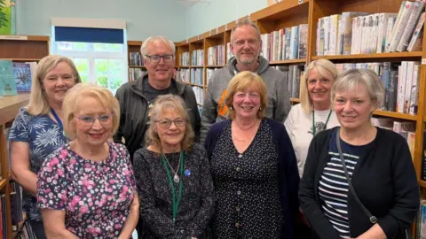 The photo shows 8 people smiling at the camera. They're standing in a library. There are shelves behind them, with books on. 