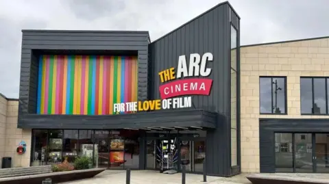 The Arc Cinema A black building frontage with The Arc Cinema logo on the side. There are a number of multi-coloured vertical panels on its left hand side.