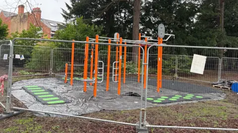 Charnwood Borough Council Bright orange fitness equipment behind fencing in a soggy-looking park
