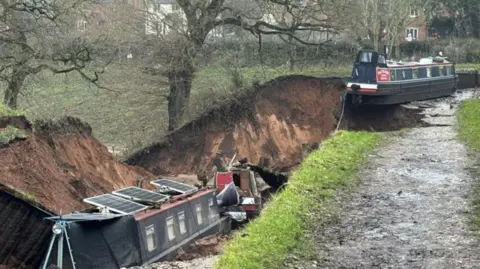 Shropshire Fire and Rescue Service A canal boat in a large muddy trench and another in the background hanging over the edge of the hole, with a wet, grass-edged footpath running alongside