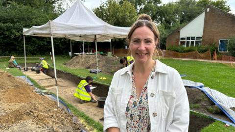 Sue Potts standing in front of the Roman road, with soil and turf dug to the side of it and four people behind her. Ms Potts is wearing a patterned shirt and white overshirt 