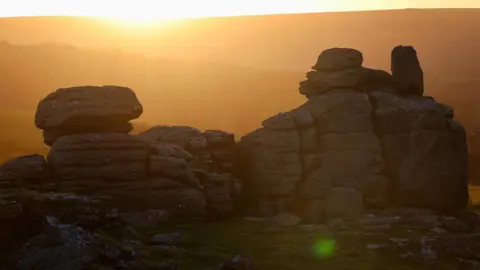 Jelly B Photography A rocky tor with an orange sun setting behind hills in the distance. 