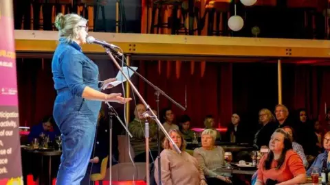 A woman standing on a stage reads words from a piece of paper into a microphone. There are people sitting on tables on the floor in front looking up at her. She is wearing a blue denim outfit and her blonde hair is tied back. The room is draped in long red curtains.