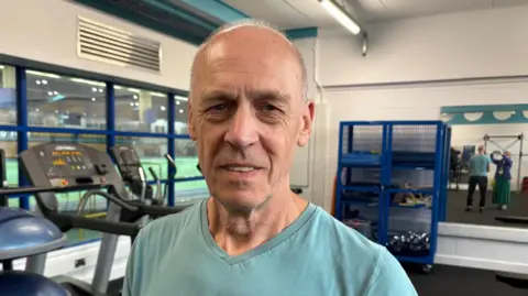 Anthony is stood in one of the gyms at Cardiff Met university, where he continues his physiotherapy since his cardiac arrest. Behind him are running machines and free weights. He is wearing a green t-shirt and looking at the camera. It is a head and shoulders shot of him
