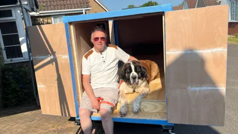 John Devine/BBC Alan Valkeith and his dog Louis, who is a brown, black and white St Bernard, are sitting in a homemade caravan which will be used by them both. It is painted blue and is sitting on a driveway.