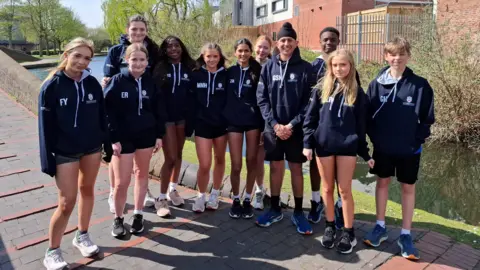 A group of students wearing sporting attire standing at the end of a bridge over a river, with a building and the water visible behind them.