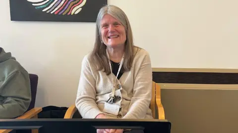 Music therapist Satyam Makoieva is pictured sitting on a chair in front of a beige-coloured wall. She has long grey hair and is wearing a beige cardigan with a black top underneath. She is also wearing a lanyard and she is smiling widely.