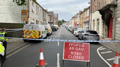 A street blocked off by police tape and cones. You can see this is the case at either end of the road. There is a sign which says FFORDD AR GAU/ Road CLOSED. Police vans line the left side and an officer is visible.
