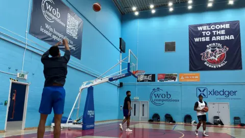 A trio of Bristol Flyers basketball players practice inside their home court in Filton. One of them is shooting a long-distance attempt at the basket while the other two watch. The walls of the sports hall are a light blue, with some sponsors names and logos written on them in black