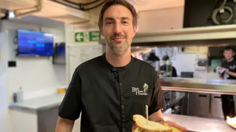Mark Ansell/BBC A chef in black uniform holds up a sandwich in a pub kitchen.