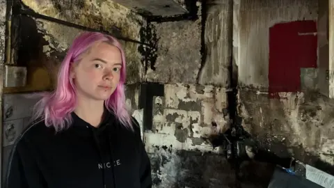 A woman with light pink hair and a black top stands in the burnt out kitchen with carred walls behind her.