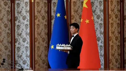 Reuters A man in a black suit and bow tie carries a tray of teacups. He's walking in front of a China flag and EU flag standing side by side.