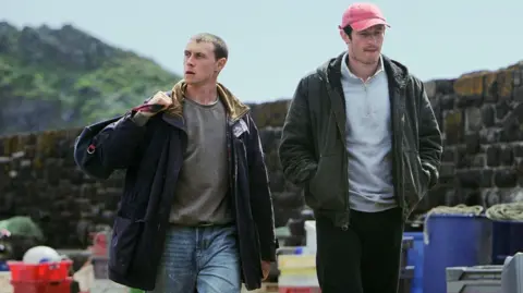 Ian Kingsnorth A scene from the film in which George MacKay and Callum Turner are walking along the harbour, MacKay has a bag slung over his shoulder. There are fishing boxes lying on the quay beside them and a low wall runs behinds them.