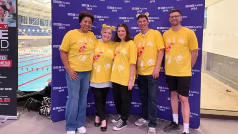 BBC Five people wear yellow t-shirts in front of a purple board with BBC Radio WM branding. The pool is visible in the background to the left.
