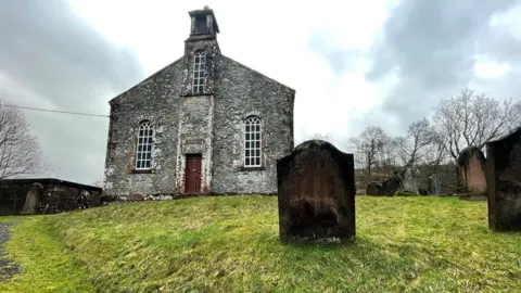 Wamphray Church is sitting on a hill in a countryside setting with headstones around it. There are gloomy skies above and some trees, without leaves, beside it.
