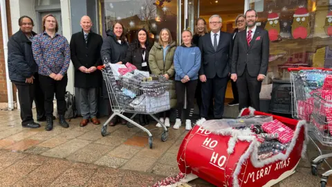 BBC A group of people stand outside a cafe and presents are packed into trollies and a small red sledge which has ho ho ho Merry Xmas written in white on it.