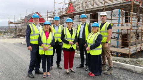 The Braidwater Group A group of seven people, all in hard hats and yellow high viz jackets stand in front of a house that is under construction. the house is covered by scaffolding.