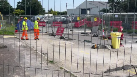 Two workmen in high vis jackets and hard hats survey their work within a fenced off area. Red signs behind the fencing read 'danger' while orange cones are also dotted around