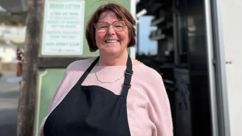 Rhona Hewit is smiling while standing in front of her kiosk. She is wearing a black apron and pink top.