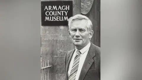 Armagh County Museum A black and white photograph of Roger, who is dressed in a suit and wearing a stripy tie. He is standing in front of a sign that says Armagh County Museum.