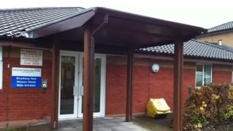 BBC The entrance of a one-storey, redbrick building with white PVC double doors. There are four pillars holding up a roof coming out from the doorway. A blue and white sign with the NHS logo on it reads 'Broadway Park, Wessex House, Main Entrance'.