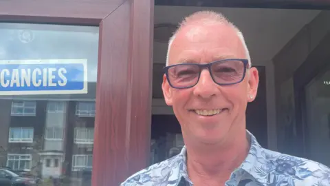 A picture of Paul Gregory-Ward taken outside the Corona Hotel in Blackpool. He is standing in front of glass UPV brown doors, one side is closed, the other open. There is a blue sign with the word vacancies in white lettering to his left stuck inside the window. Paul is wearing a floral shirt, with blue glasses and an earring in his right ear. He has balding grey hair and is smiling.
