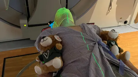 Anthony Perriam Anthony Perriam is lying in a hospital bad with two teddy bears by his side. He is wearing a mask as he is about to be scanned for cancer.