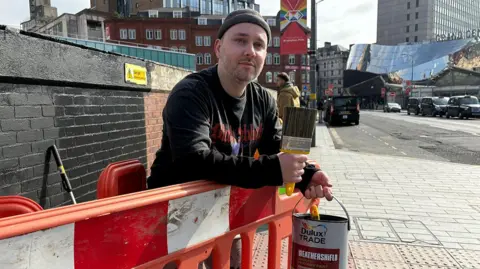 A man stands whilst leaning over an orange barricade. He is holding a yellow paintbrush in one hand and a pot of Dulux paint in the other. Behind him a brick wall is being painted black. Cars, taxis and the top of Grand Central station can be seen in the background behind him.