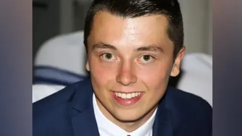 Connor Brown looking directly into the camera and smiling. He has dark hair, brushed to his right, and is wearing a navy suit, white shirt and red tie.