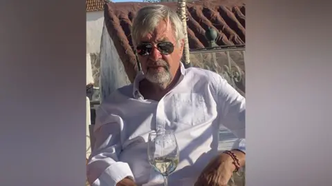 Suffolk Police A man with grey hair wearing a white shirt sits at an outdoor table with a glass of white wine. He wears sunglasses along with a bracelet on one wrist.
