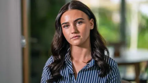 A young women with long wavy dark hair stares expressionless at the camera, wearing a blue and white striped shirt.