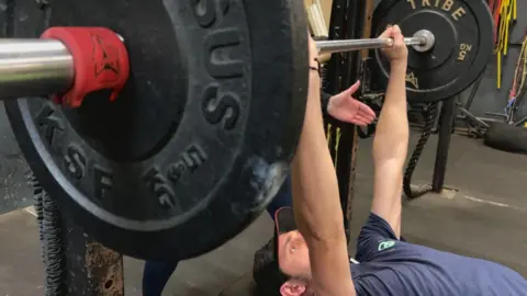 A man with a blue T shirt and black trousers and a dark cap holds a barbell above his head while lying on a bench.