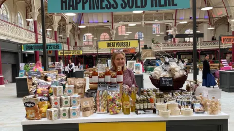 A smiling woman in a red top standing behind the counter of a market stall.