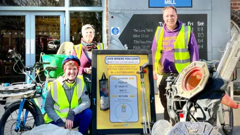 Stroud District Council A group of three people - two women with short hair and a man, all middle-aged - stand around a yellow A-board, which has litter pickers and bin bags stuck to it.