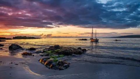 A coastal sunset, with a boat resting in the bay as water laps around it, and dramatic clouds overhead and a sky coloured purple, blue and orange. 