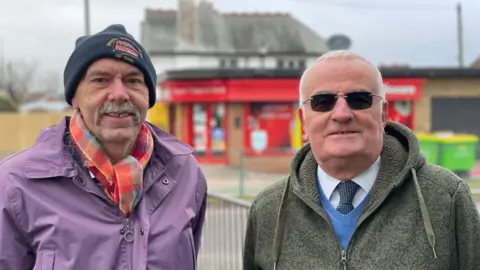 Two men standing outside. On the left the man has a grey moustache and stubble, and is wearing a navy beanie hat, red and blue scarf and a purple waterproof jacket. The man on the right has short white hair and black tinted glasses on. He wears a white shirt with a navy and white tie, blue jumper and dark green hoodie.