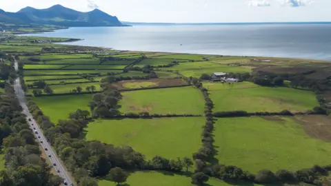 Carter Jonas Multiple green fields surrounded by hedgerows and trees can be seen right along a section of the coast, with farm buildings and smaller buildings seen in the distance. Mountains can also be seen in the distance.