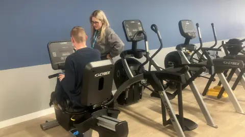 Photograph of the gym in the Ged Mason building at the Seashell Trust. The image shows a student on an exercise bike, with one of the teachers standing next to him. 