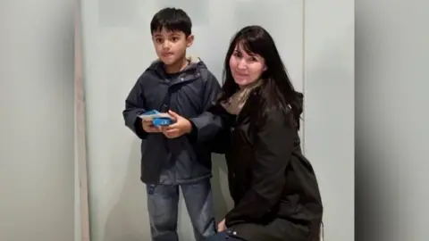 Family handout A young boy, with short brown hair, wearing denim jeans and a navy coat. A woman with long black hair is kneeling beside him. She is wearing a cream scarf and a black coat. 