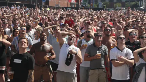 Getty Images Hundreds of England football supporters stand looking at a big screen as England play. Many of them have their hands on their heads as though a big chance has just been missed. It is a sunny day and some fans have their tops off