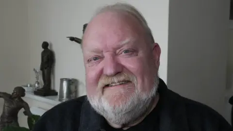 John Fairhall/BBC A head and shoulders image of Colin Kreidewolf. He is wearing a black top and smiling while looking into the camera. He is sitting in front of a light-coloured wall and there are statues behind him.