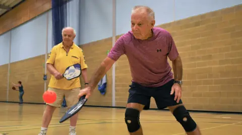 Two men aged over 55 are engaged in a game of indoor pickleball. The men are both wearing shorts and a T-shirts.