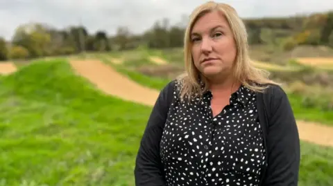 Angela Ruskin wearing a black polka dot shirt and black cardigan. She has blonde hair and is looking sternly at the camera.