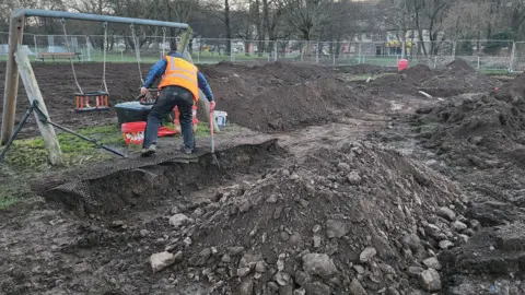 Mark Mather A children's park has been completely dug up with numerous piles of earth and metal fencing all around the site. Just one swing remains standing, a man is digging in front of it. 
