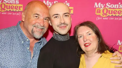 Getty Images A person with a bald head is standing with a bald man with grey facial hair and a woman with brown hair. They are close together and have their arms around each other, and are standing in front of a pink backdrop with a logo on that reads "My Son's A Queer but what can you do?"